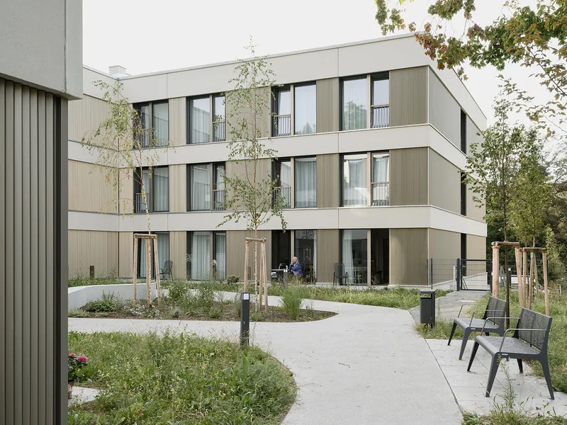 Courtyard pathway winding through young trees and planted beds toward the stepped residential volumes