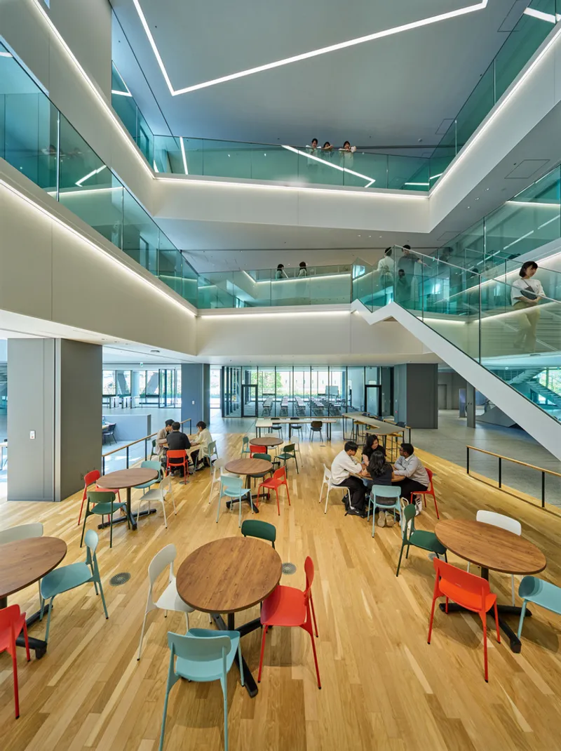 Timber-floored cafeteria with round tables overlooked by stacked glass balconies with integrated LED edge lighting