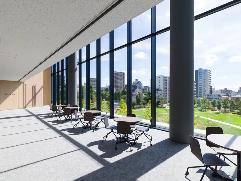 Corridor with small round tables beneath tall black-framed windows overlooking a green plaza in afternoon sunlight