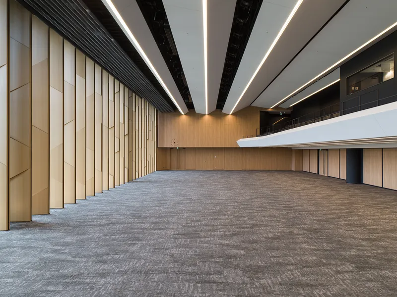 Large assembly hall with vertical timber paneling and radiating linear ceiling lighting above carpeted floor
