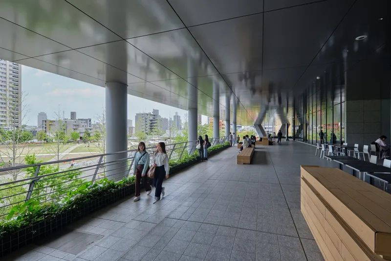 Open-air corridor with planted edge and visitors walking beneath angled white columns on an overcast day