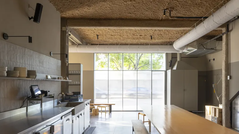 Interior with exposed hempcrete ceiling, stainless steel counter, and floor-to-ceiling glazing filtering daylight onto wooden tables