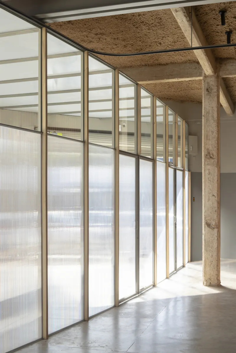 Curved translucent glass partition with brass frames beneath an exposed hempcrete ceiling and concrete column