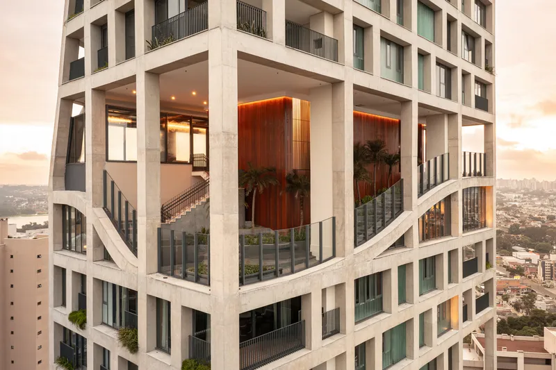 Corner view of the raised concrete podium with diagonal bracing, glazed terrace, and timber volume at dusk