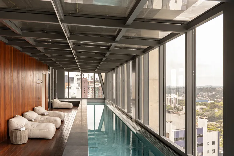 Indoor pool with exposed steel ceiling grid, floor-to-ceiling glazing, and timber wall under overcast sky