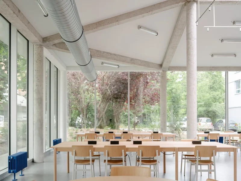 Dining area with timber furniture beneath exposed concrete beams and circular ductwork facing courtyard trees