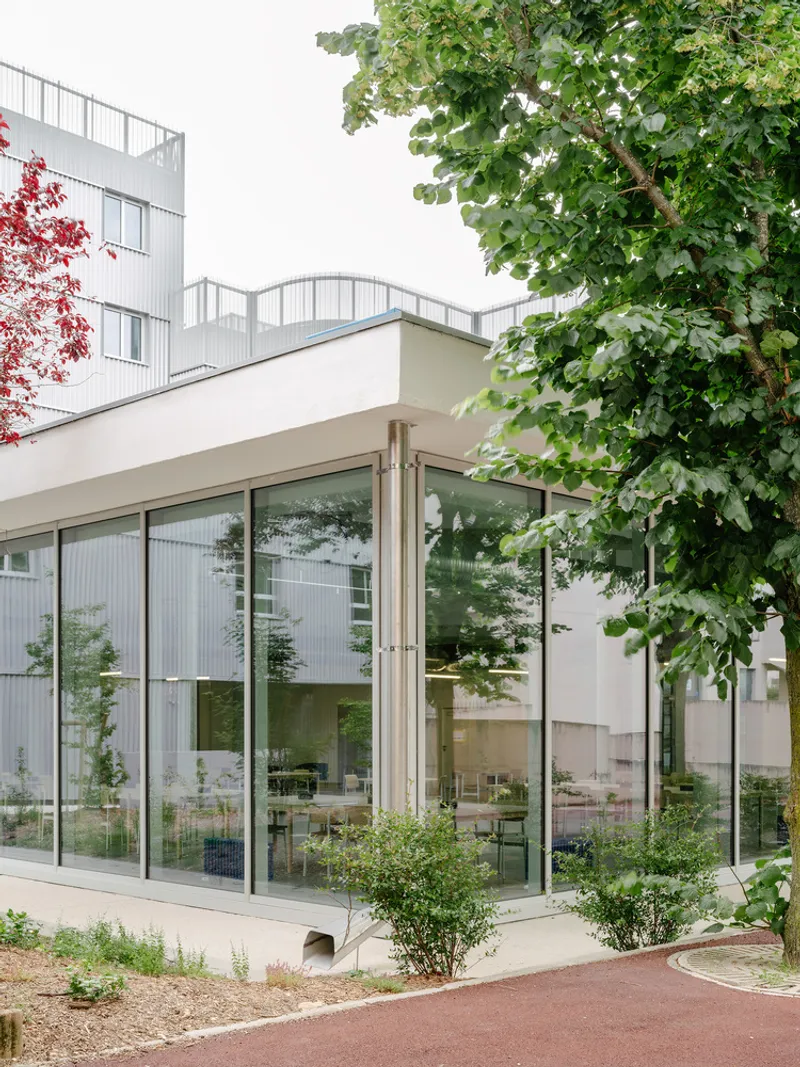 Corner glazed pavilion with flat roof surrounded by planted beds and trees beneath residential buildings
