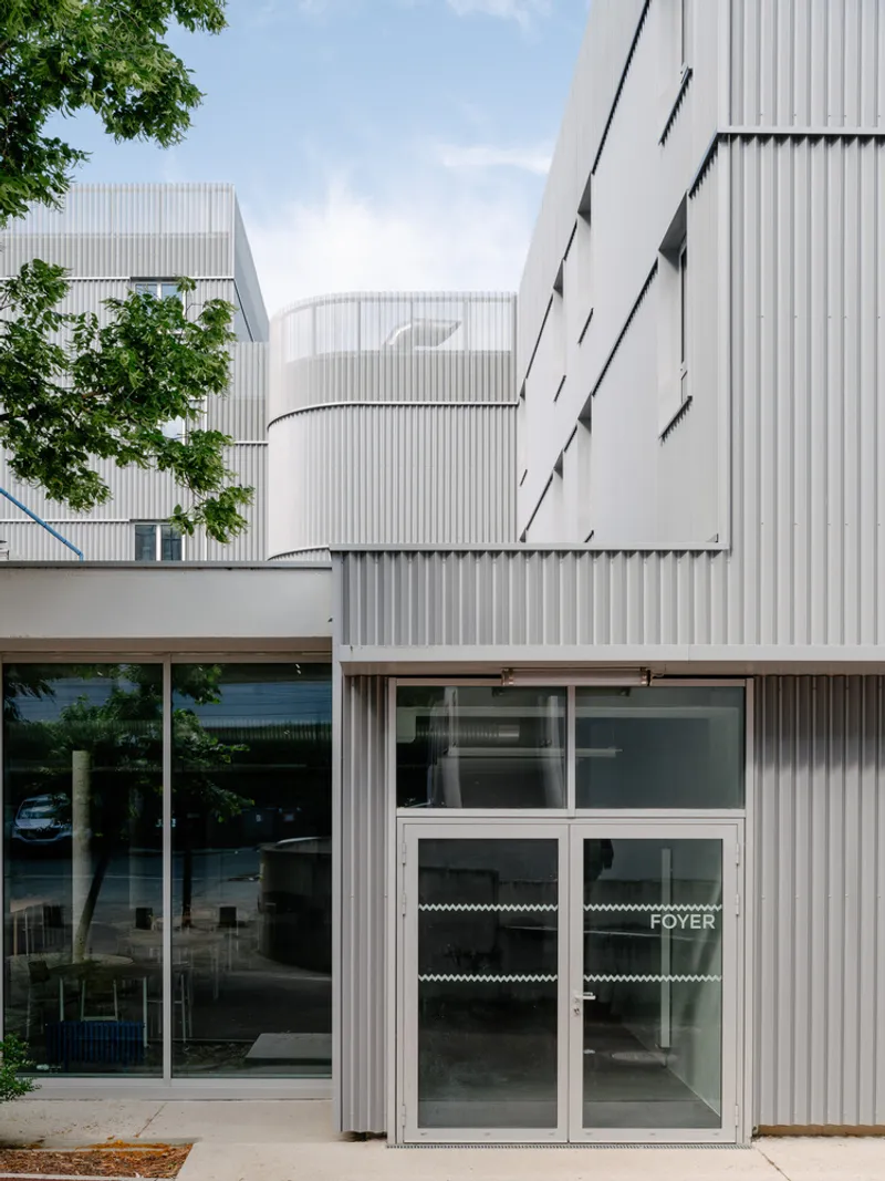 Ground-level entrance with glass doors and corrugated metal cladding framed by tree branches overhead