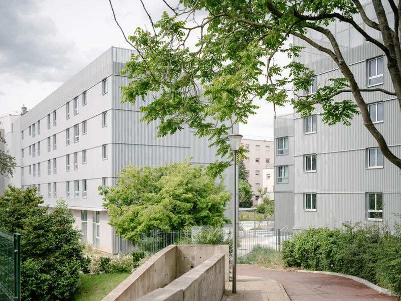 White corrugated metal facade of a multi-storey residential block framed by mature trees under an overcast sky