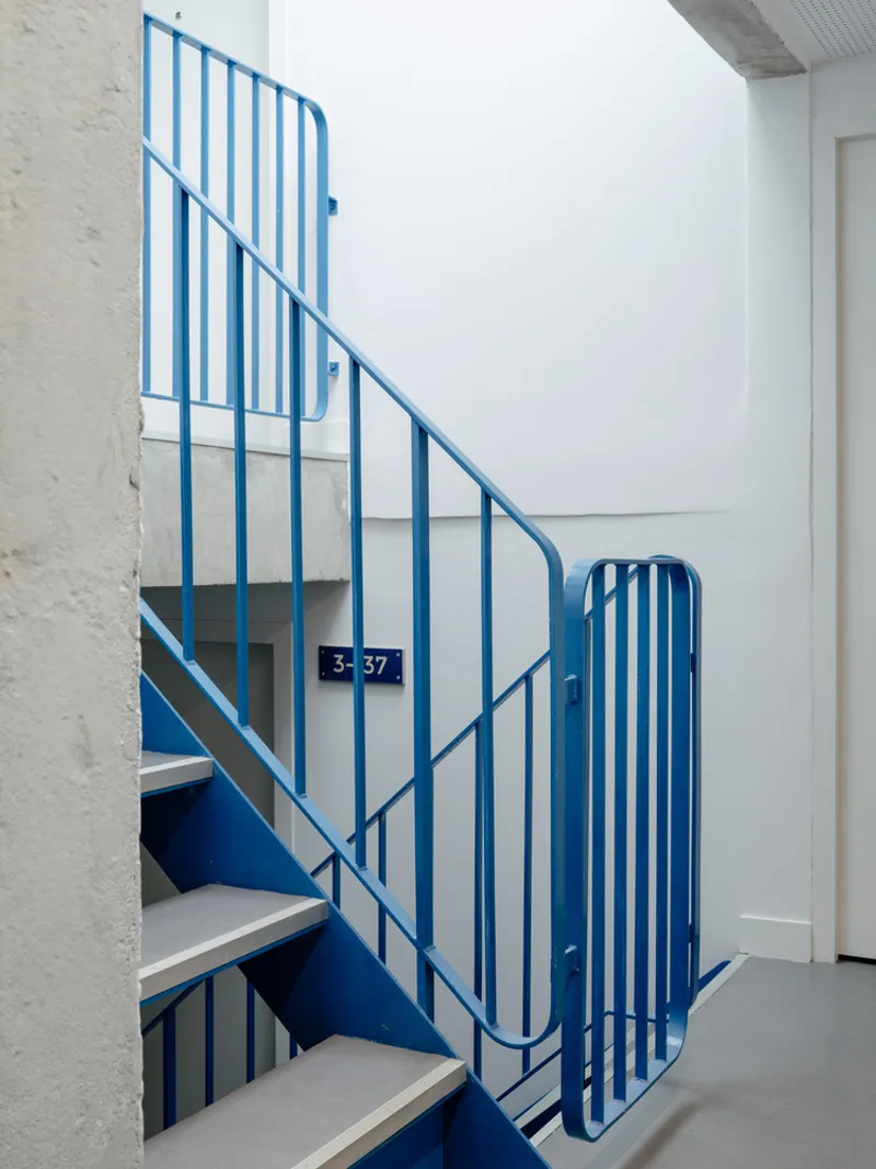 Blue steel staircase with vertical bar railings and unit number plaque in a white-walled corridor