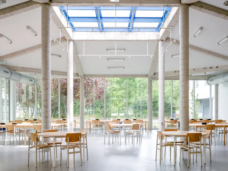 Communal dining hall with timber tables and chairs under exposed concrete beams and blue-framed skylight