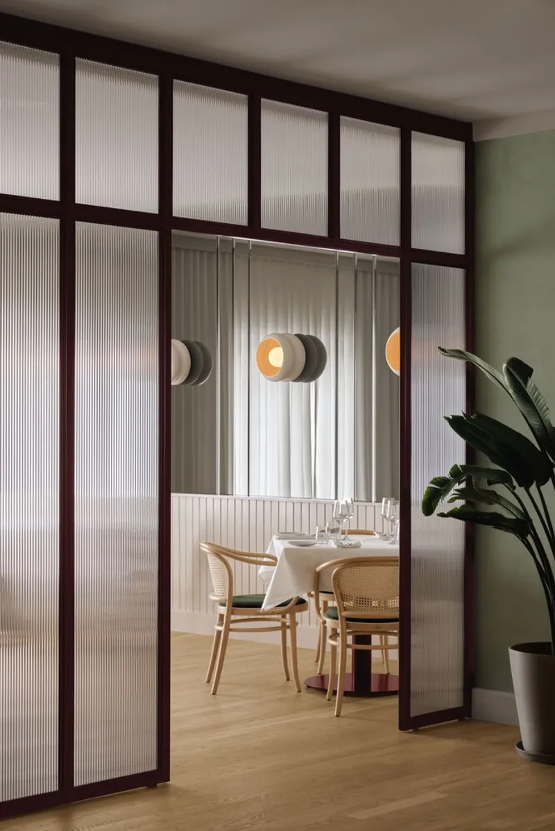 Fluted glass partition with dark timber frame separating two dining spaces, potted plant at right