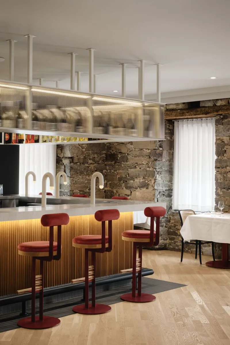 Bar counter with red velvet stools beneath a suspended glass rack and exposed stone wall