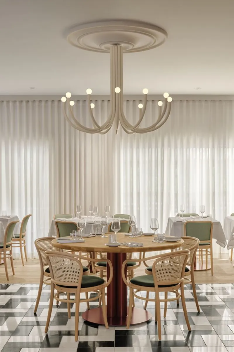 Round table with cane chairs beneath a chandelier, checkered floor tiles and sheer curtains behind