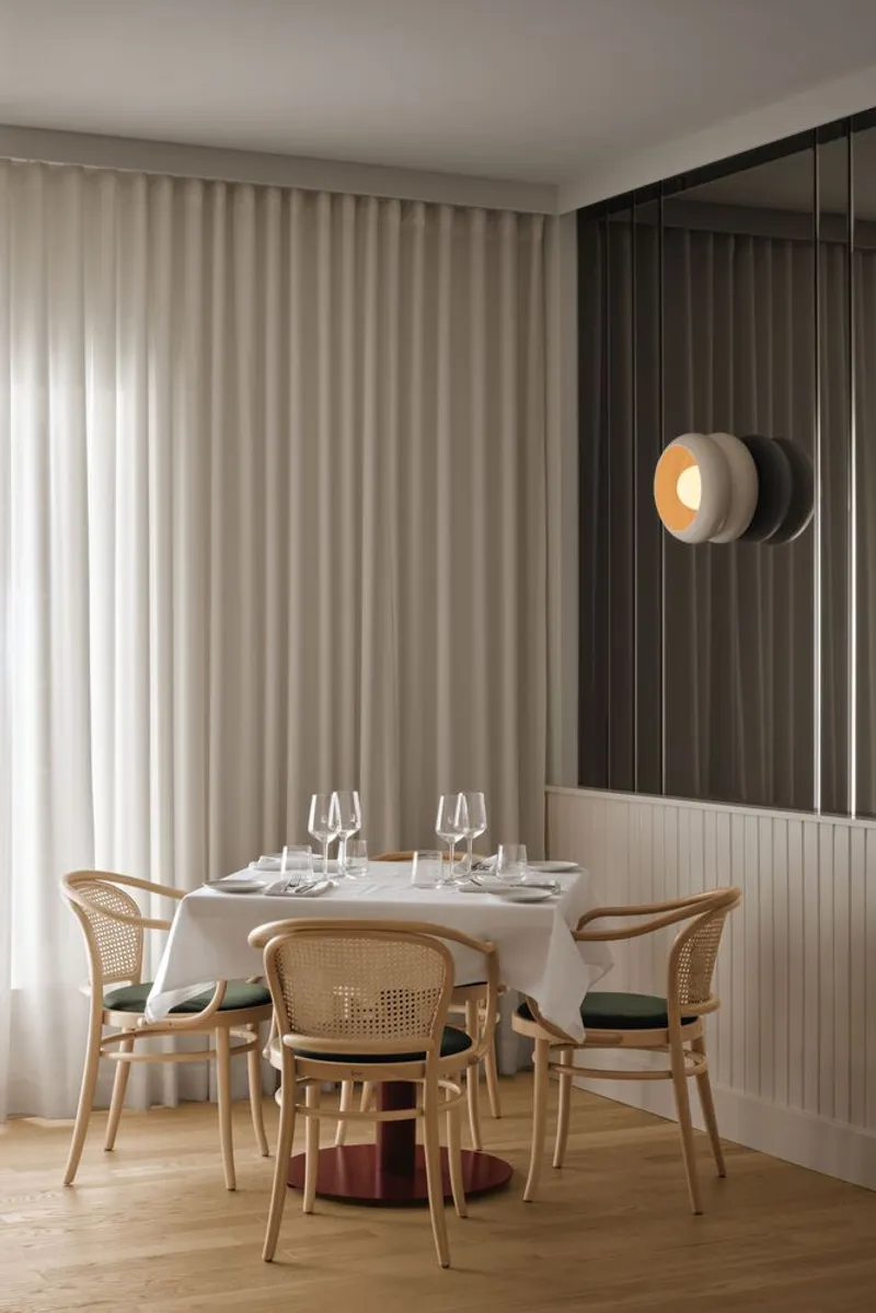Dining table set with cane chairs beneath floor-to-ceiling curtains and mirrored fluted wall panels