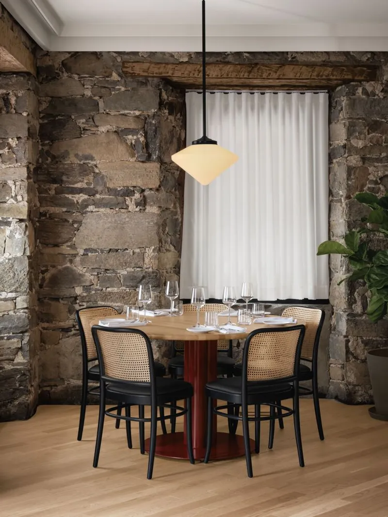 Dining table with cane chairs beneath pendant light against exposed stone wall with timber beams