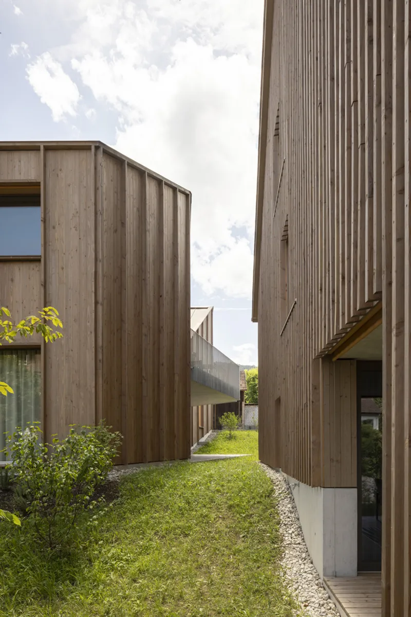 Passage between two vertical timber-clad volumes with grass underfoot and distant lawn beyond