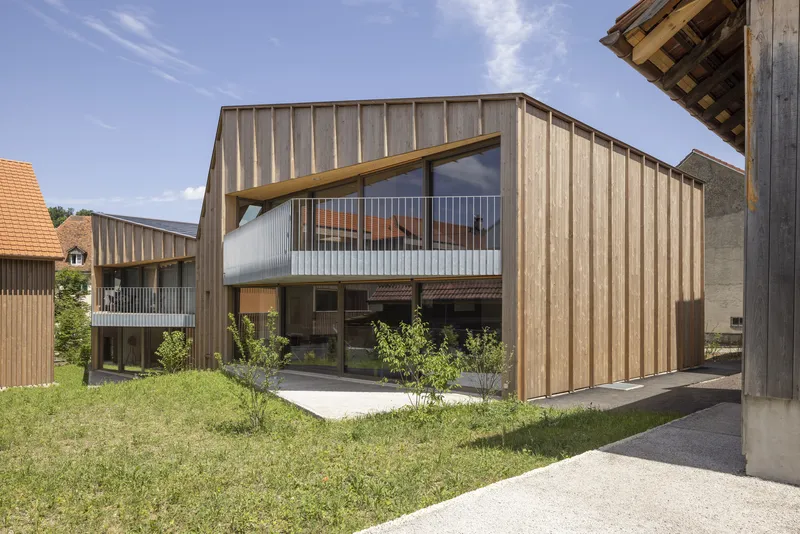 Gabled timber facade with translucent balcony screen overlooking gravel and newly planted trees