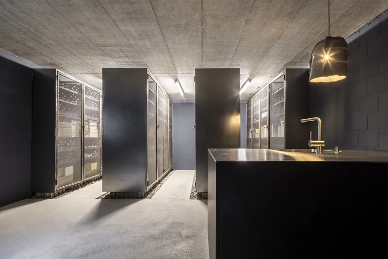 Wine cellar with exposed concrete ceiling, black metal partitions, glass-fronted storage racks, and pendant light above island