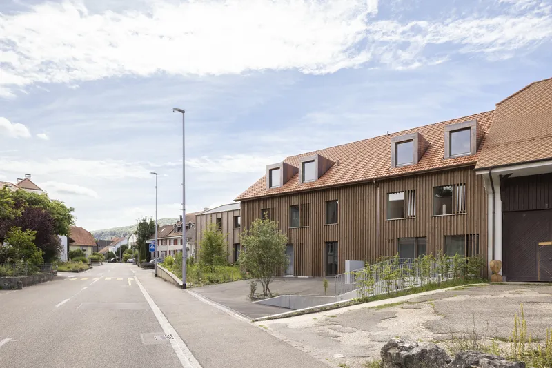Street view of vertical timber-clad facade with dormer windows beneath orange clay tile roof on partly cloudy day