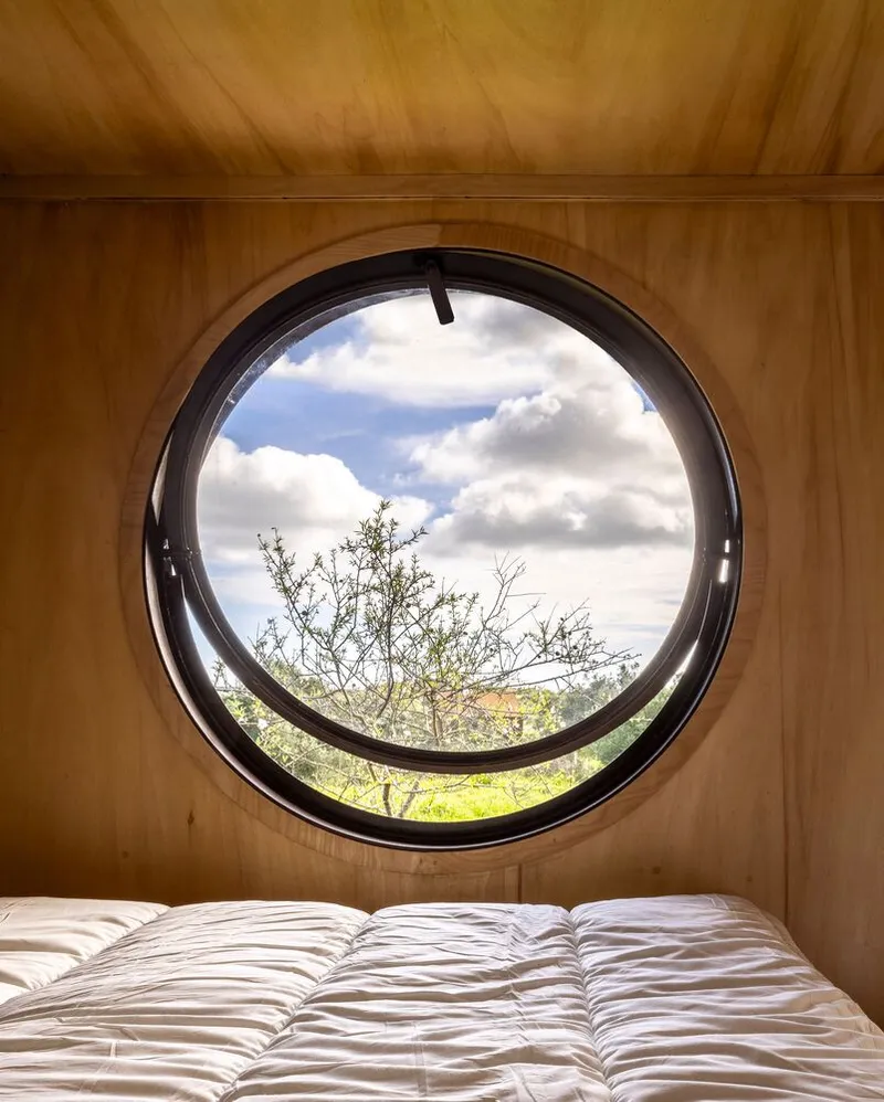 Circular porthole window framing trees and cloudy sky above a bed with white quilted bedding