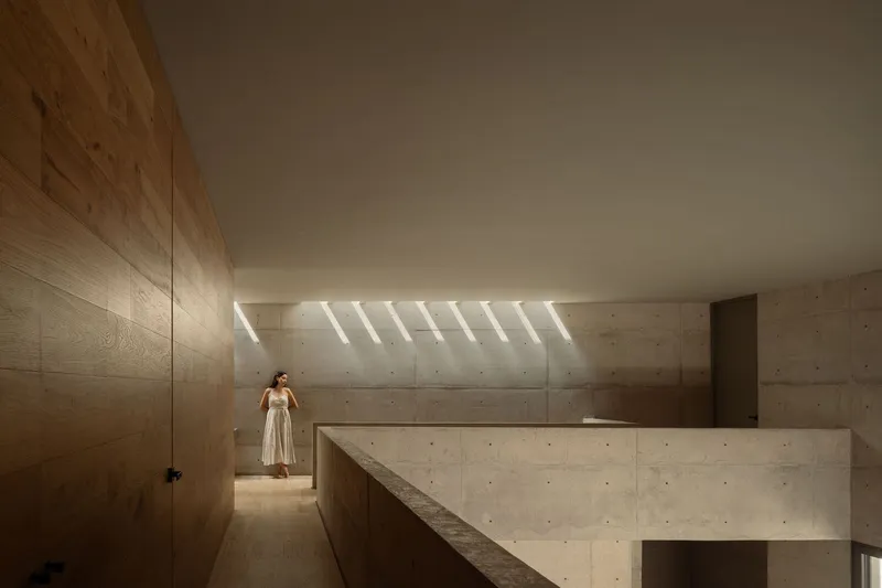 Interior hallway with board-formed concrete walls and wood paneling illuminated by clerestory skylights with a figure standing