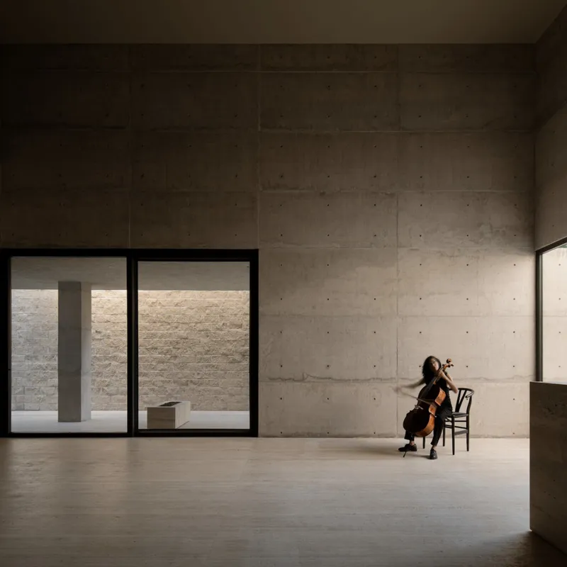 Board-formed concrete interior with cellist seated by floor-to-ceiling glazing overlooking a courtyard