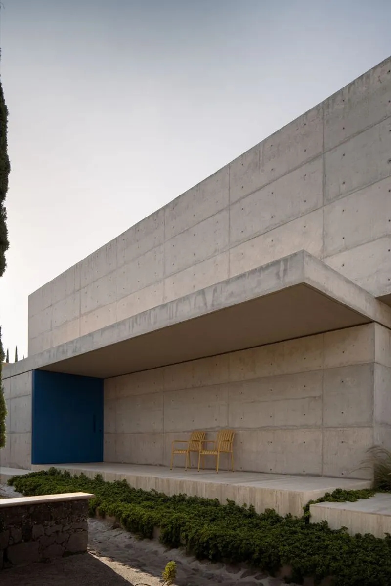 Cantilevered concrete overhang sheltering a recessed porch with two timber chairs and low planting