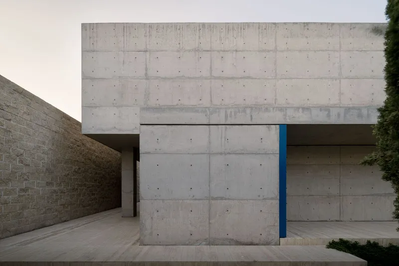 Entrance courtyard framed by board-formed concrete walls with a recessed blue door under overcast light