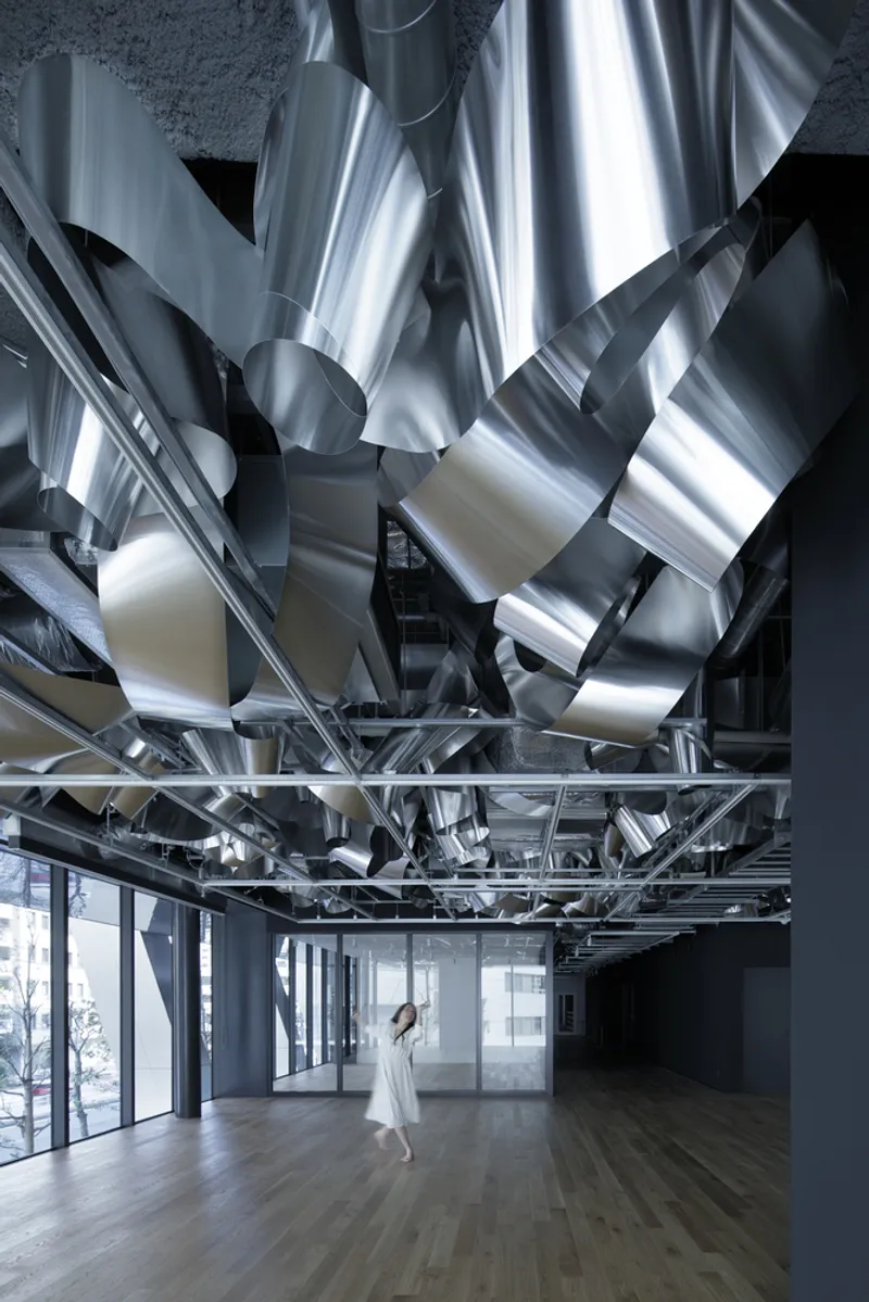 Person standing in a daylit corridor below the cascading sculptural metallic ceiling and steel support grid