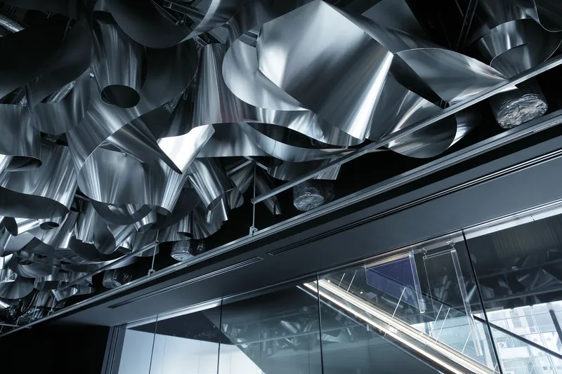 Polished stainless steel sculptural ceiling installation above an escalator and glass curtain wall