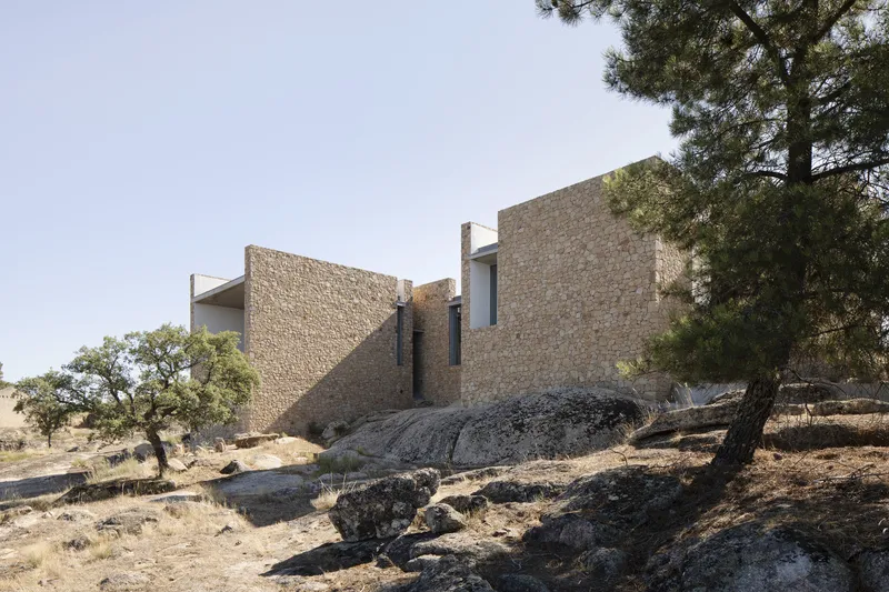 Stone-clad volumes rising from a rocky hillside terrain with scattered pine trees and dry grasses