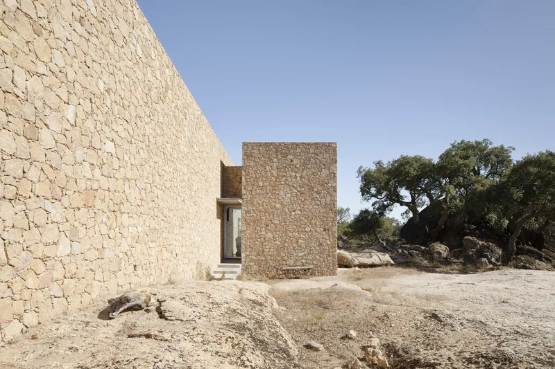 Stone facade volumes framing an entry passage with ancient olive trees and rocky terrain beyond
