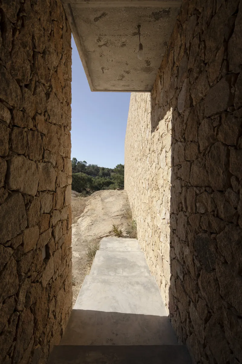 Concrete-lined passageway framed by stone walls opening to a sunlit hillside with scattered vegetation