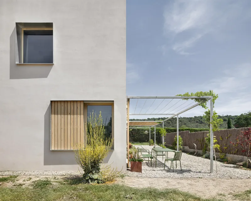 Exterior courtyard with white render facade, timber shutters and cable-supported shade canopy under blue sky