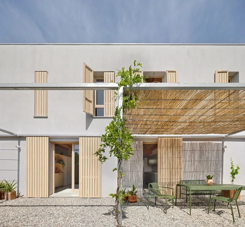 White stucco courtyard facade with vertical timber shutters and a woven reed pergola over green metal lounge chairs