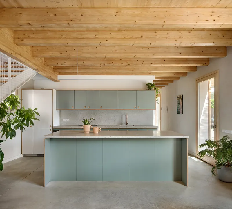 Kitchen island with sage green cabinetry and terrazzo countertop beneath exposed timber joists and floating timber stair