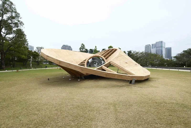 Tilted timber pavilion with circular opening in a park with trees and distant towers