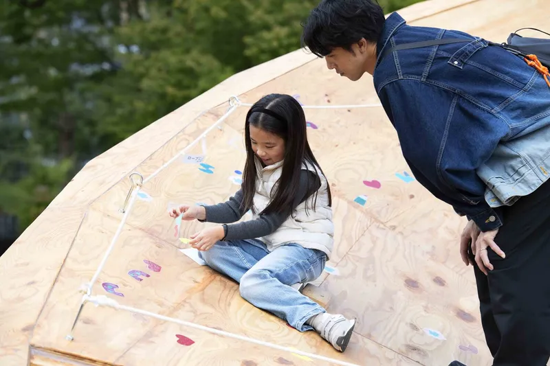 Child drawing with chalk on the sloped plywood surface while an adult watches