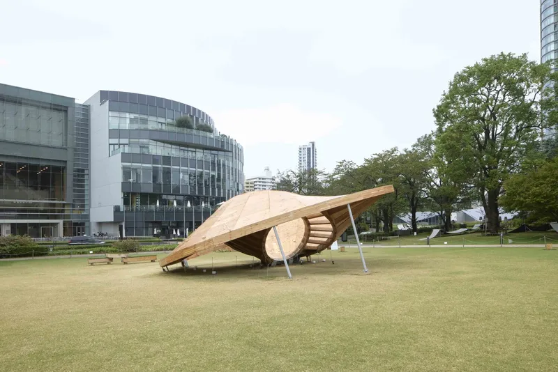 Sculptural timber pavilion with sweeping shell form on a green lawn beside glass buildings