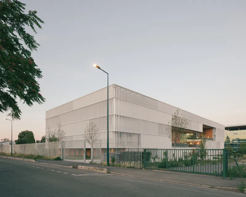 Street view of the louvered white volume behind metal fencing and a lit streetlamp at dusk