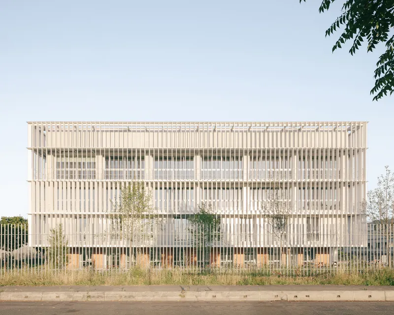 Street view of the four-story facade with vertical timber louvres and young trees behind a metal fence