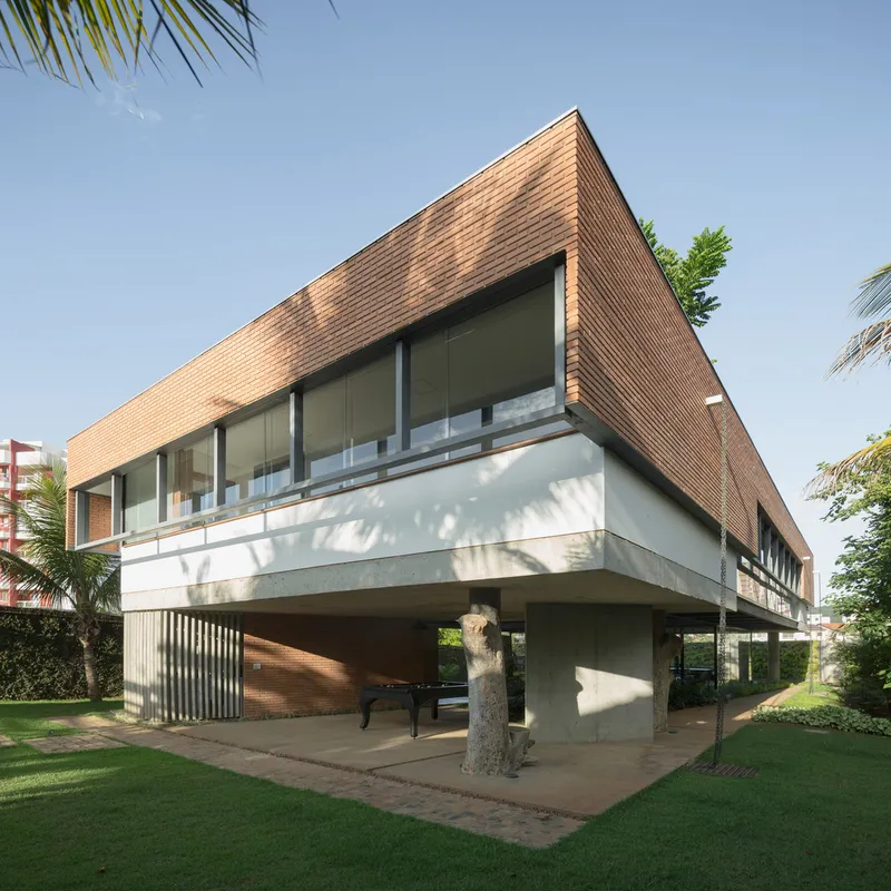 Cantilevered upper volume with angled wood cladding supported on concrete piers in sunlit lawn with palms