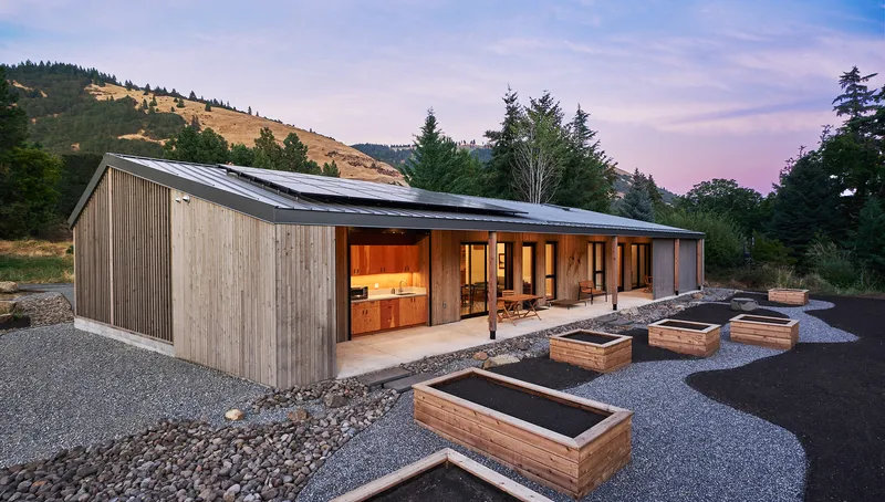 Vertical timber clad volume with metal roof and glazed openings beside raised garden beds at dusk