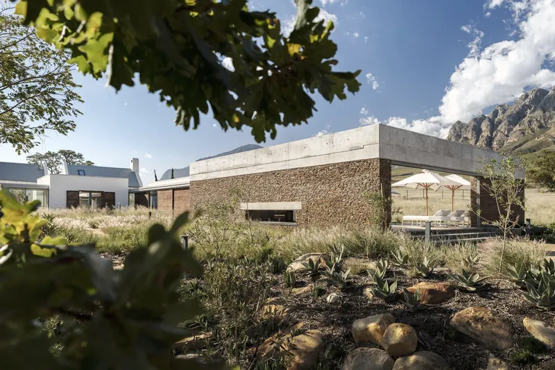 Concrete-framed volume with stacked stone cladding viewed through native plantings and oak leaves