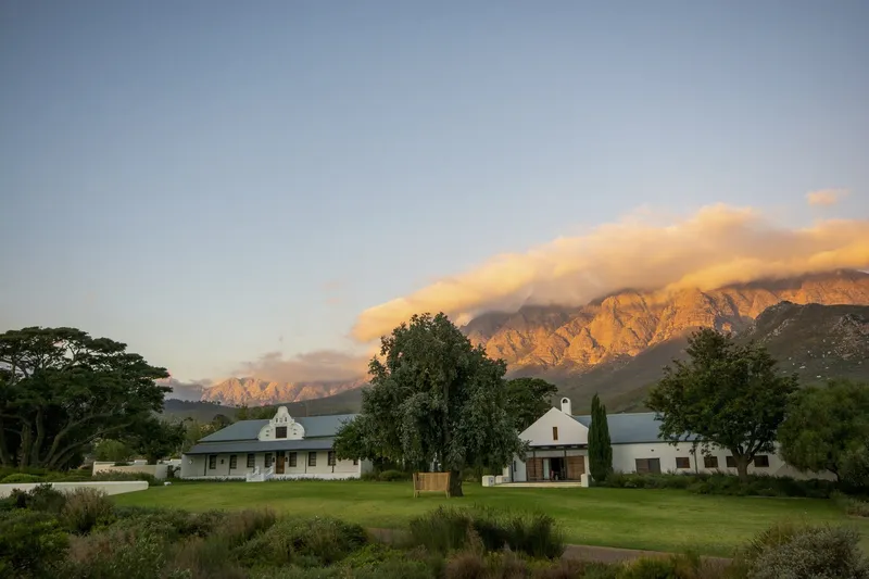 Long view of white gabled buildings set in a lawn with mountains at sunset