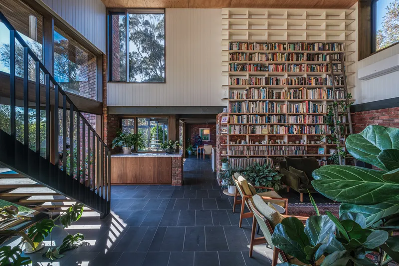 Double-height living space with floor-to-ceiling bookshelf wall, black steel staircase and indoor plants on dark tile floor