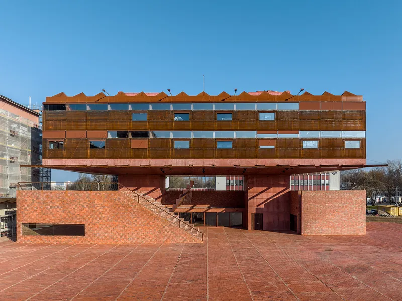 Elevated corten steel volume on red concrete base with perforated metal screens and brick plaza under clear blue sky