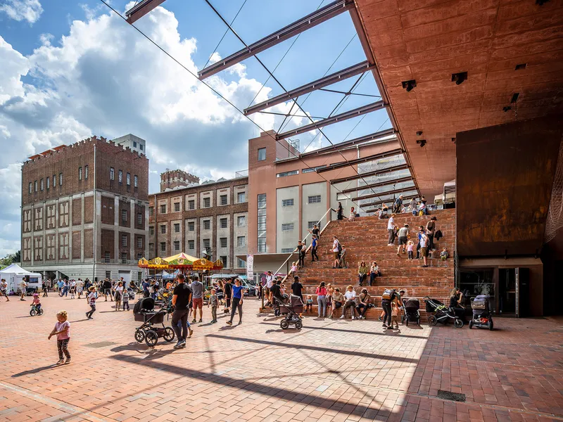 Public plaza with brick paving and open steel truss canopy overhead filled with visitors on a sunny day