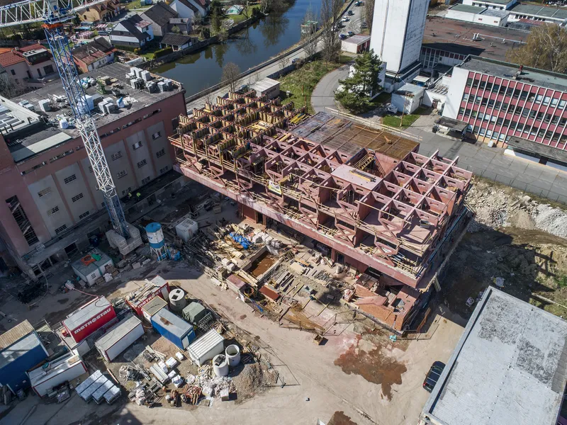 Aerial view of construction site showing corten steel frame structure with timber formwork beside a canal and industrial buildings